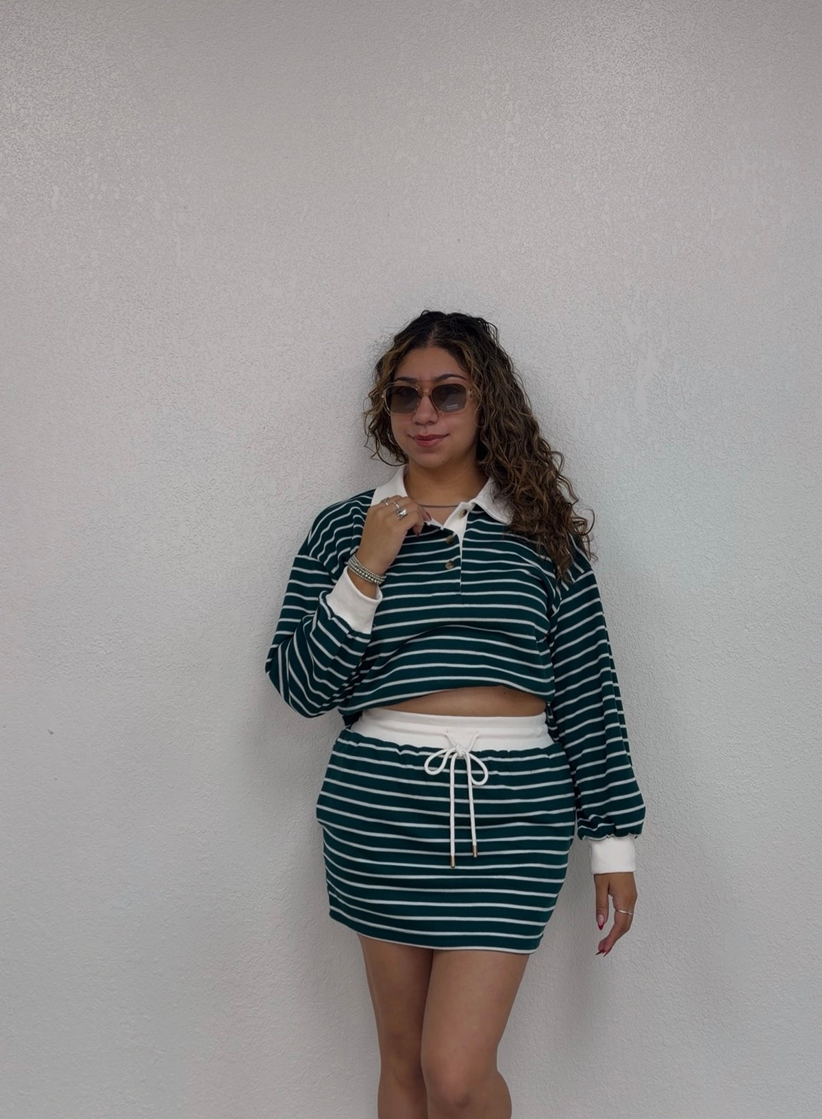 A woman modeling a matching two-piece set consisting of a dark green and white horizontally striped long-sleeve crop top with a white collar and a matching striped mini skirt with a white drawstring, standing against a textured off-white wall.