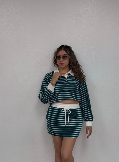 A woman modeling a matching two-piece set consisting of a dark green and white horizontally striped long-sleeve crop top with a white collar and a matching striped mini skirt with a white drawstring, standing against a textured off-white wall.